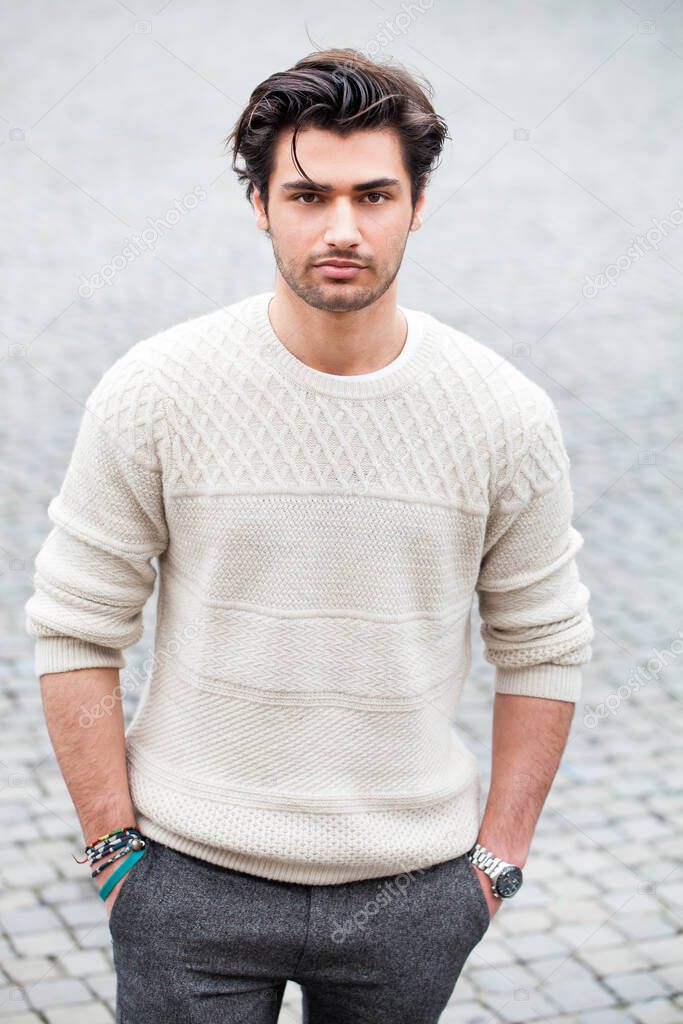 Handsome young man outdoor in the street with hands in the pockets. Fashion hairstyle, confident attitude. White sweater. Stubble.