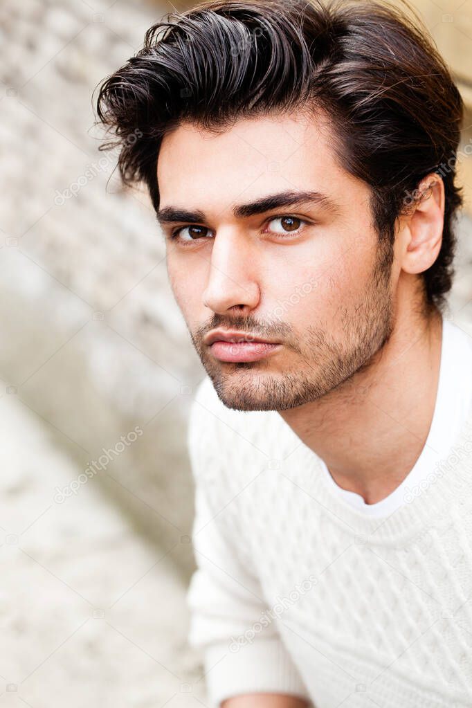 Beautiful young man outdoor. Close portrait, fashion hairstyle. Charming and attractive look. Stubble and white sweater. Outdoors in the city.