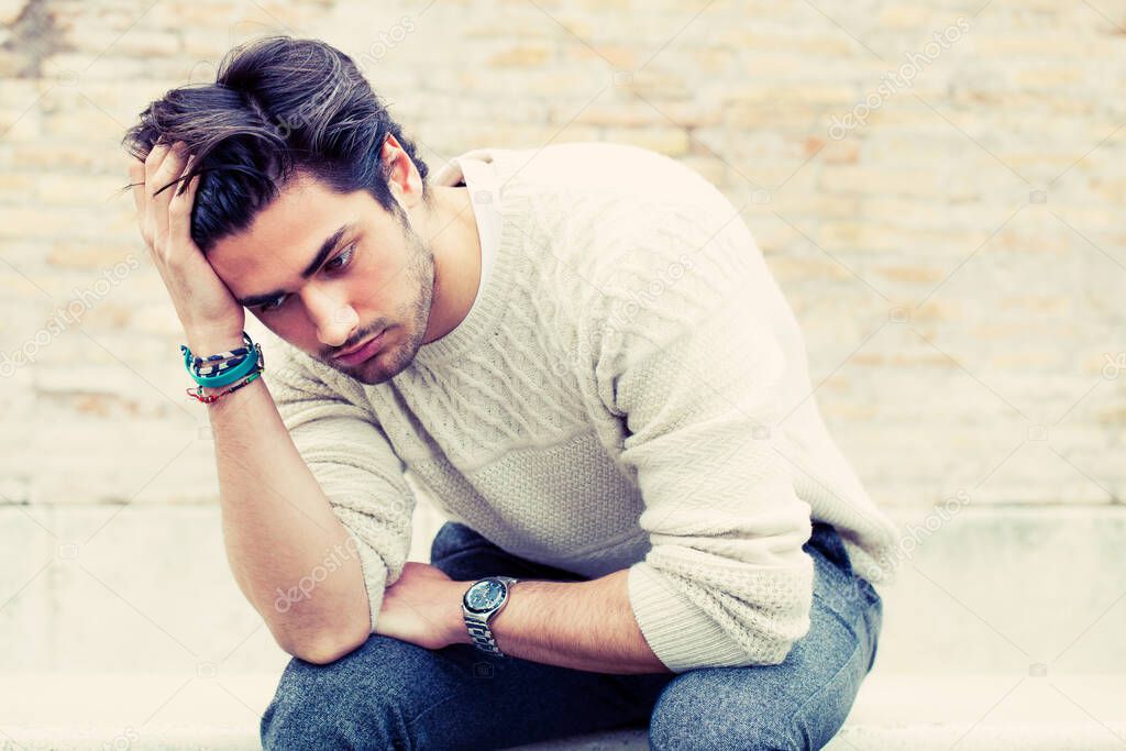 Young worried man sitting and staring outdoors with hands on the head and hair with desperate attitude. White sweater. In the street.