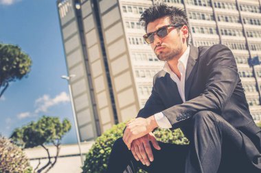 Dışarıda oturan güneş gözlüklü yakışıklı adam modeli. Arkasında ofisleri olan bir bina var..