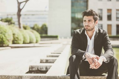 Şehirde oturan yakışıklı genç bir adam. Kolları bacaklarındayken rahat bir düşünce. Siyah ceketli ve pantolonlu bir takım elbise..