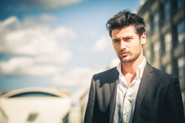 Takım elbiseli ve beyaz gömlekli adam. Şık saçlı ve sakallı. Şehrin caddesinde açık havada. Gökyüzü ve arkaplanda binalar.