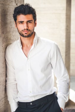 Portrait of young handsome man in white shirt outdoor. Nice appearance with stylish hair and beard. Leaning with a side on a wall clipart