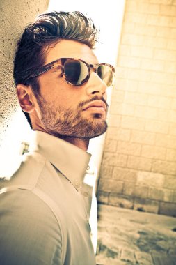 Dışarıda güneş gözlüklü havalı ve yakışıklı bir adam. Duvara yaslanarak. Yumuşak sakallı, çekici ve çekici bir görünüş. Beyaz gömlek. Yoğun sıcak ışıklar. Yukarı bakıyorum..