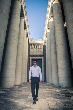 Tarihi bir binanın antik sütunlarında yürüyen modaya uyan genç bir adam. Beyaz gömlek ve siyah pantolon. Moda saç ve sakal.