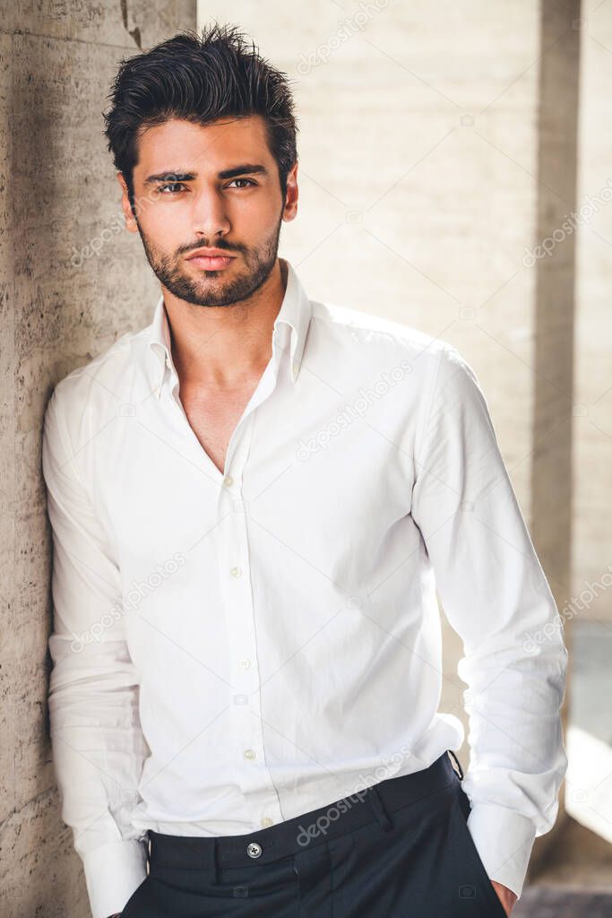 Portrait of young handsome man in white shirt outdoor. Nice appearance with stylish hair and beard. Leaning with a side on a wall