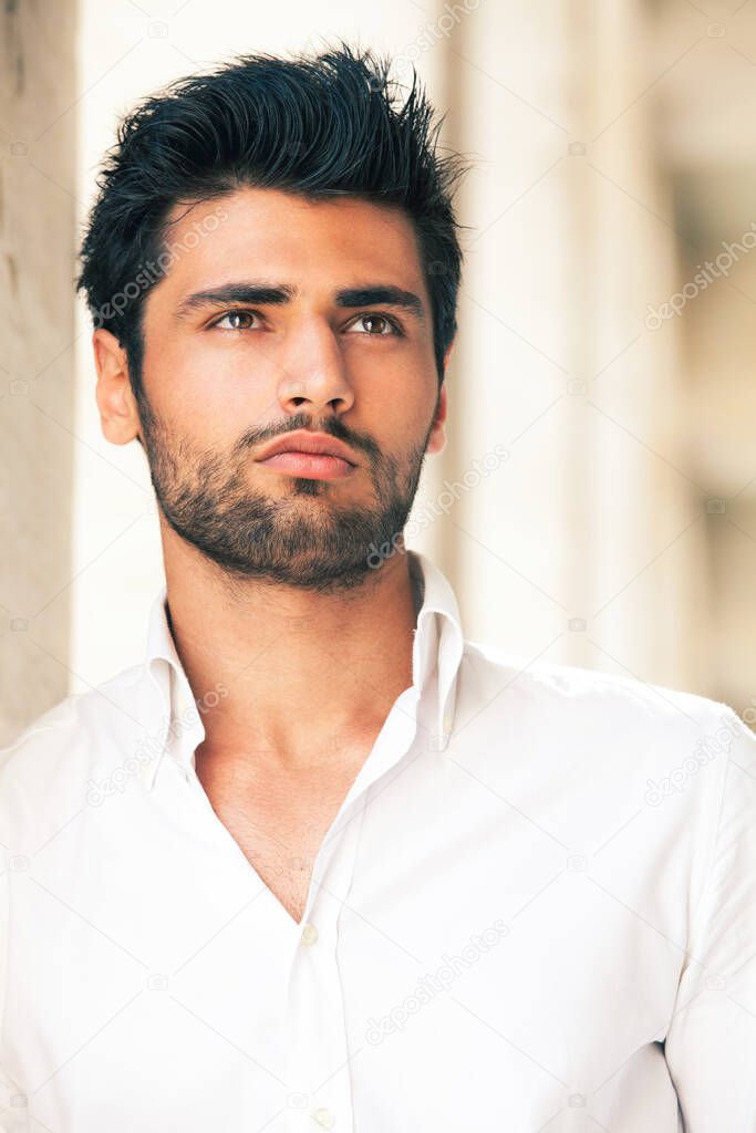 Handsome and stylish young man. Hair and beard fashionable. A beautiful young Italian man is outdoors. He is leaning against a marble wall. He wears a white shirt. Hair and black beard.