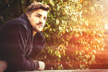 Arkasında yapraklar ve ağaçlar olan bir parkta bekleyen yakışıklı genç adam. Genç adamın hafif bir sakalı var. O bir ceket giyiyor..