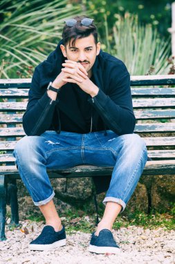 Kürsüde oturan yakışıklı genç adam modeli. Yakışıklı genç bir adam bankta oturup izliyor ve bekliyor. Moda kıyafetleri giyen adam. Yanındaki boş bankta..
