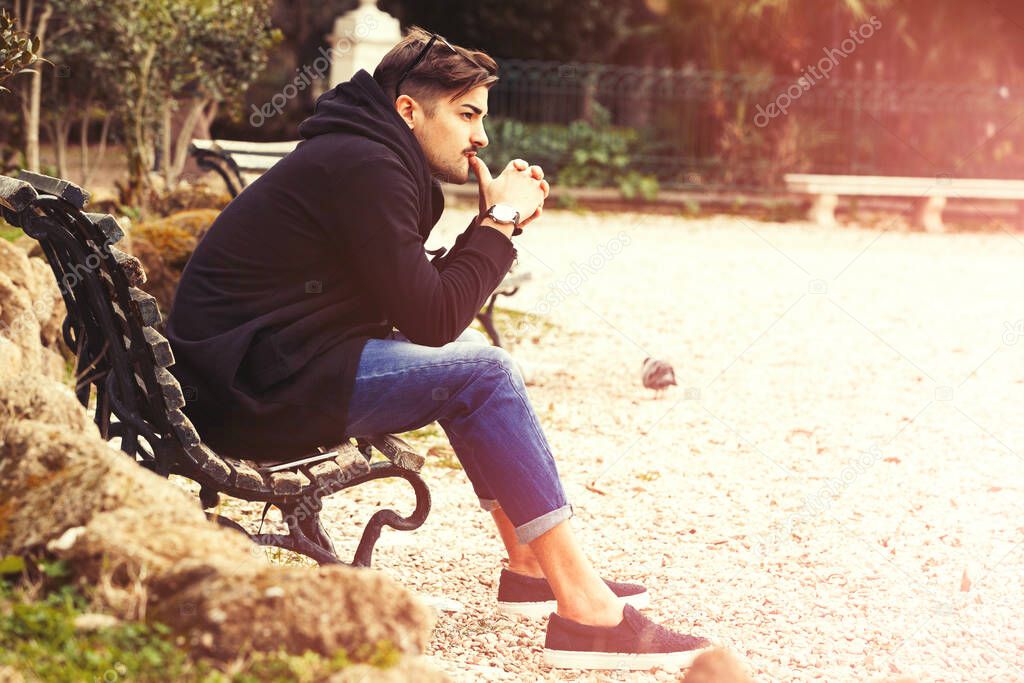 Handsome young man model sitting on the bench. A handsome young man is sitting on a bench watching and waiting. The guy wearing trendy clothes. Beside him the empty bench.