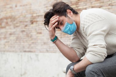 Covid19, endişeli genç adam oturuyor ve yere bakıyor. Dışarıda, elleri kafasında, hüzünlü bir tavırla. Coronavirus kişisel koruma ekipmanları ppi.