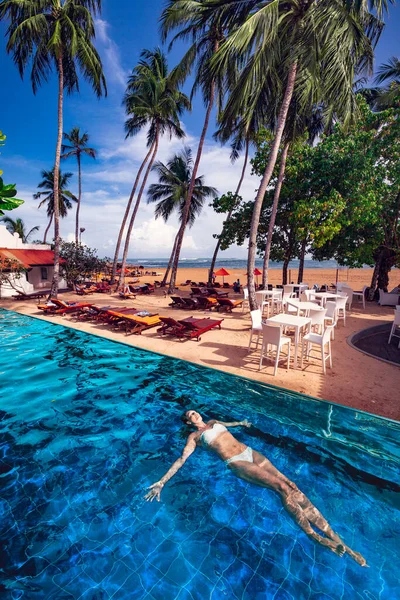 Woman swimming floating in the pool in a tropical seaside resort on the beach. Deck chairs and umbrellas, palm trees and sky. Sea holidays. — Stock Image Woman Swimming Floating Pool Tropical Seaside Resort Beach Deck Chairs Royalty Free Stock Images