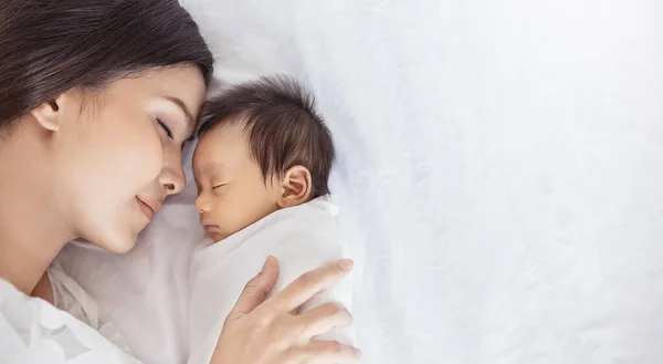 近景肖像美丽的年轻亚洲或高加索母亲女孩亲吻她的健康新生儿睡在床上与复制空间 亚洲妇女的生活方式母亲节观念 图库照片