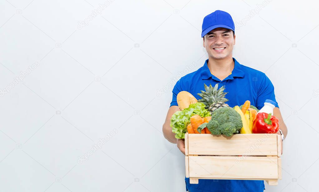Closeup portrait of beautiful caucasian delivery man in blue uniform holding wood basket of grocery fresh vegetables food isolated on white. Healthy lifestyle shopping internet online supermarket delivery concept.
