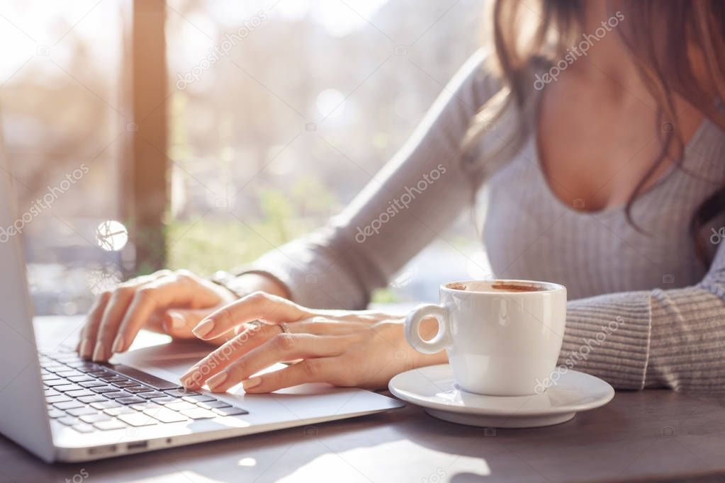 A girl works behind a laptop in a cafe, fingers typing on the keyboard. Workplace in a cafe, a cup of coffee next to a laptop, toned bright sunny photo