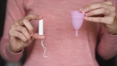 Elinde farklı türde hijyen ürünleri tutan bir kadın. Kadın adet bardağını seçiyor ve tampon atıyor.