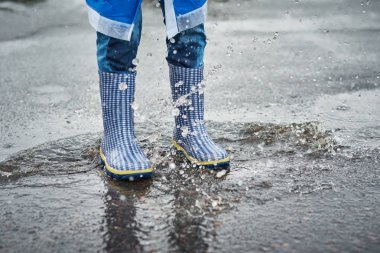 Mavi çizmeli çocuk yağmurda su birikintisinin üzerinden atlıyor.