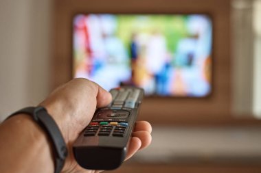 Odada televizyon izleyen ve remo kullanarak kanalları değiştiren bir adam var.