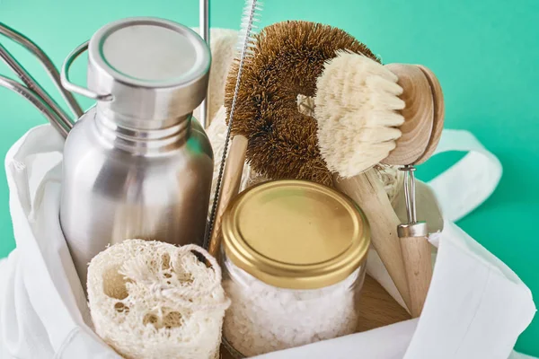 Reusable plastic free items in cotton shopping bag. Glass jar, metal straws, aluminum bottle and wooden cleaning brush on a green background — Stock Photo, Image