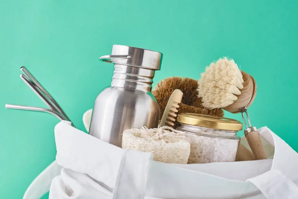 Reusable plastic free items in cotton shopping bag. Glass jar, metal straws, aluminum bottle and wooden cleaning brush on a green background — Stock Photo, Image