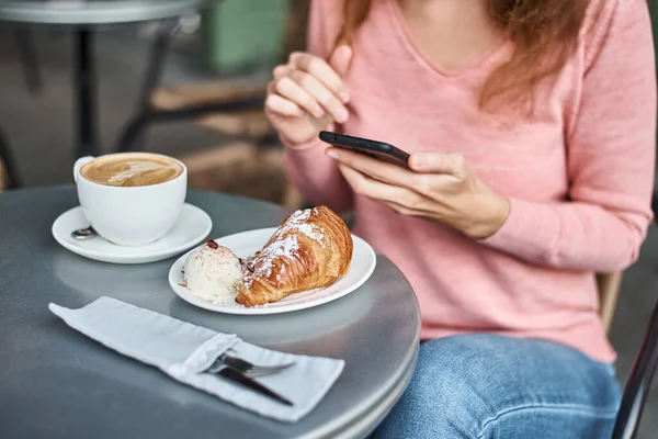 Woman have breakfast in caffe and using smartphone. Girl chatting and using internet with phone during coffee break with croissant