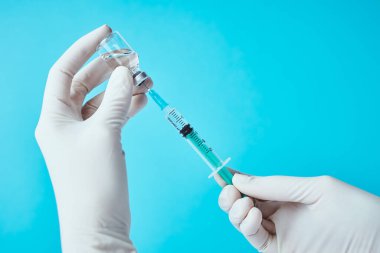 Eldivendeki eller mavi arka planda şırınga ve ilaç ampulü tutuyor. Sağlık, tıbbi ve virüs koruma kavramı