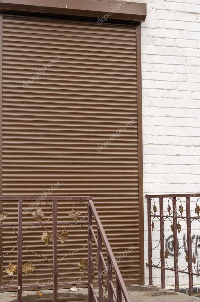 The main entrance is closed with a protective shutter. Door in a white brick wall with anti-vandal protection. The entrance is equipped with forged railing. Selective focus.