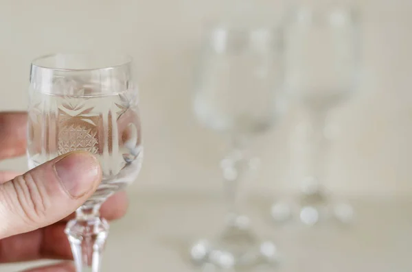 Crystal glass of vodka in the hand. The left hand of an adult man holds a full refined glass with strong alcohol. Eye level shooting. Selective focus. Blurred background.