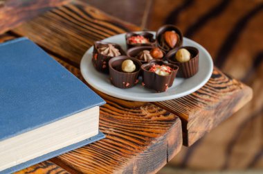 Çikolatalı şekerler beyaz bir tabakta. Kapalı bir kitabın yanındaki ahşap bir masada lezzetli şekerlemeler. Şekerleme. Yan görüş. Seçici odak.