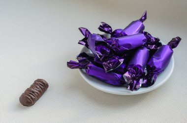 Bir fincan tabağında leylak ambalajında çikolata yığını. Lezzetli şekerlerle dolu beyaz porselen tabaklar. Yakınlarda ambalajsız bir şeker var. Göz hizasında atış. Yakın plan. Seçici odak.
