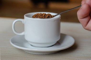 Bir fincanın üzerinde bir kaşık hazır kahve. Bir el bir kaşık dolusu hazır kahveyi beyaz bir fincana döker. Geleneksel sabah kafeinli içkisi. Yan görüş. Seçici odaklanma. Yakın plan..