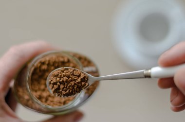 Elinde kahve olan cam kavanoz. Bir el bir kaşık dolusu hazır kahveyi tutar. Cam kavanozun üzerinde metal kaşıkta hazır kahve taneleri yığını. Kafein içeceği. Yukarıdan bak. Seçici odaklanma. Yakın plan..