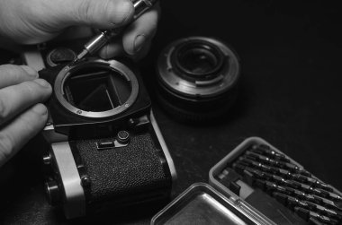 Siyah bir masada parçalanmış eski model bir kamera. Eski bir analog kamerayı tamir et. Elim merceklerin vidalarını söküyor ve tornavidayla vidalıyor. Yan görüş. Seçici odak.