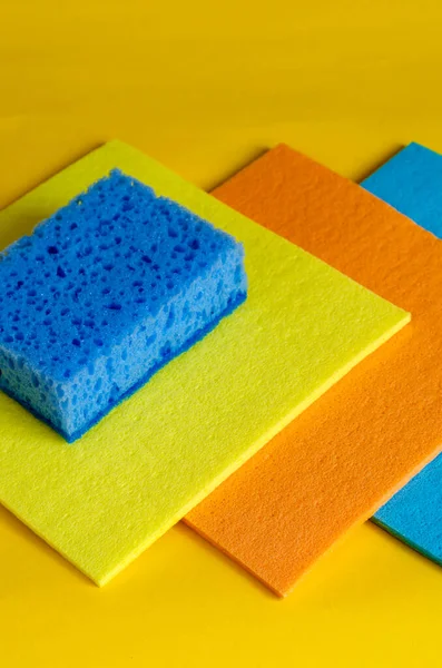 Kitchen napkins for cleaning and a sponge on a yellow background. Three multi-colored napkins for dishes and a blue porous kitchen sponge. Products for cleaning. Selective focus.