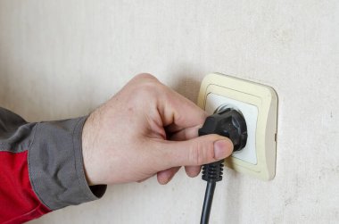 Elektrik prizinin 15. bölümünün tamiri. İş kıyafetleri içindeki bir adamın elinde prizde elektrik prizi var. Profesyonel bir elektrikçinin ticari hizmetleri. Seçici odak.