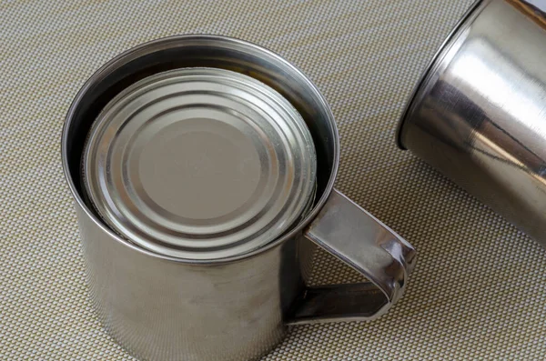 Two metal mugs and a can of canned food on a textured background. A can of canned food in a stainless steel mug. Hiking, fishing, hunting, camping. Selective focus.