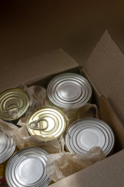 Teneke kutularla dolu açık bir karton kutu. Sabah ışınları teneke kutularda çeşitli konserve yiyecekleri aydınlatır. Gerçek konserve yiyecekler. Bir açıdan üstten bak. Seçici odak.