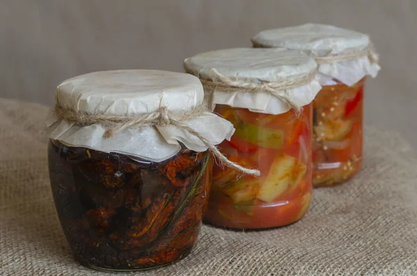 Canned vegetables, lecho and sun-dried tomatoes on burlap. Canned food in glass jars. Canning and storage of products in the cellar. Close-up.