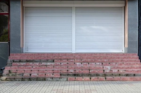 Closed shutters wide entrance to the store. Steps at the entrance with fallen off ceramic tiles. The financial crisis, bankruptcy. Front view. Without people.