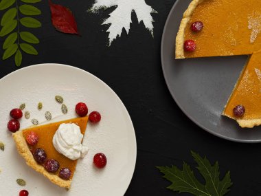 Beyaz tabakta krema ve kızılcıkla süslenmiş ev yapımı geleneksel Amerikan balkabağı turtası. Üst Manzara. Siyah arka planda renkli sonbahar yaprakları ve gri tabakta koca bir pasta var..