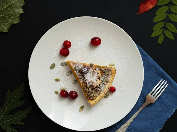 A slice of homemade traditional American pumpkin pie decorated with icing sugar and cranberries is located on a white plate. Top view. On a black background colorful autumn leaves. — Stock Photo, Image