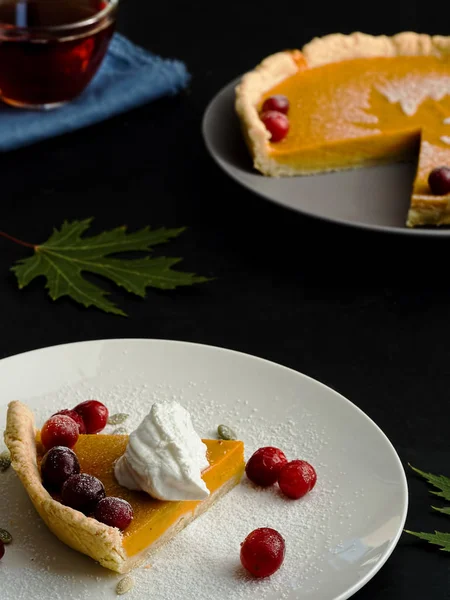 A slice of homemade traditional American pumpkin pie decorated with cream and cranberries is located on a white plate. Close-up. On a black background multi-colored autumn leaves and a cup with tea. — Stock Photo, Image