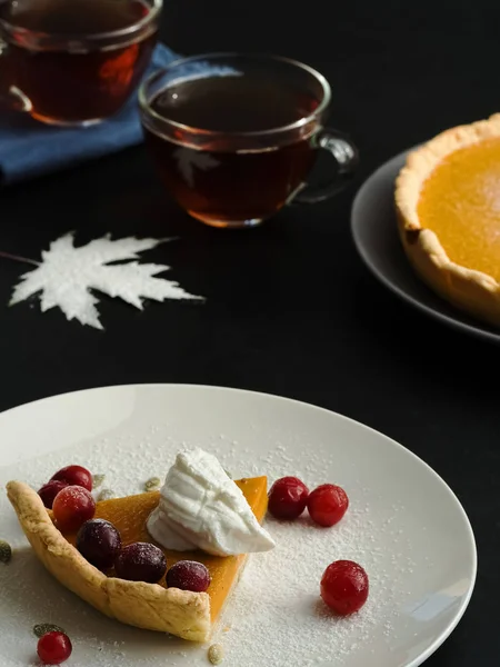 A slice of homemade traditional American pumpkin pie decorated with cream and cranberries is located on a white plate. Close-up. On a black background multi-colored autumn leaves and a cups with tea. — Stock Photo, Image