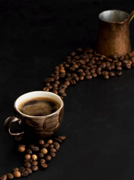 Taza con espresso oscuro sobre fondo negro. Los granos de café tostados se encuentran alrededor de una taza de café. En el fondo hay un cezve de cobre. Primer plano . — Foto de Stock