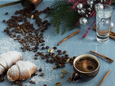 Kahverengi bir fincan espresso, kruvasan ve mavi bir fonda bir bardak su. Yakınlarda baharatlar, tarçın ve kakule ve süslemeli kozalaklı ağaçtan bir dal vardır. Üst görünüm.