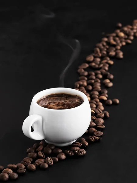 Copa blanca con espresso fragante sobre un fondo negro, vapor se eleva por encima de la taza. Los granos de café tostados se encuentran alrededor de una taza de café y en el fondo. Primer plano . — Foto de Stock