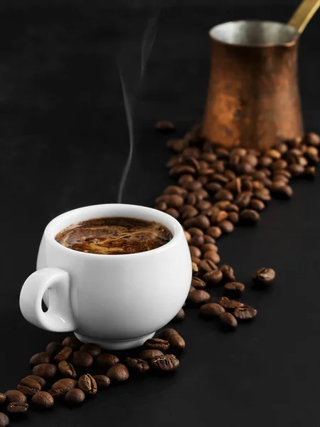 Copa blanca con espresso sobre fondo negro, vapor se eleva por encima de la taza. Los granos de café tostados se encuentran alrededor de una taza de café y en el fondo. En el fondo hay un cezve de cobre. Primer plano . —  Fotos de Stock