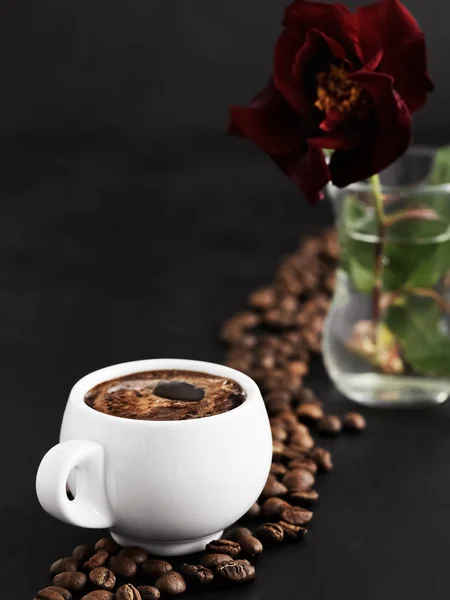 Taza blanca con un espresso fragante sobre fondo negro. Los granos de café tostados se encuentran alrededor de una taza de café y en el fondo. Primer plano. Rosa roja en un vaso sobre un fondo — Foto de Stock