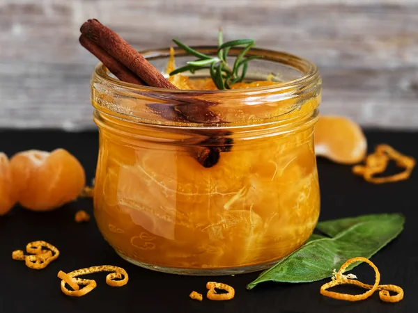 Citrus (tangerine) jam in a glass jar on a table with a cinnamon stick and a branch of rosemary. Around segments of mandarin, shavings of zest. Close-up, selective focus. Black stone background.