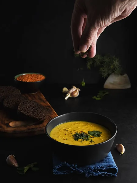 Red lentil soup, spices, herbs, vegetables and cream. Soup on a blue napkin, next to brown bread, garlic, a bowl with lentils. Female hand adds spices to the soup. Close-up.